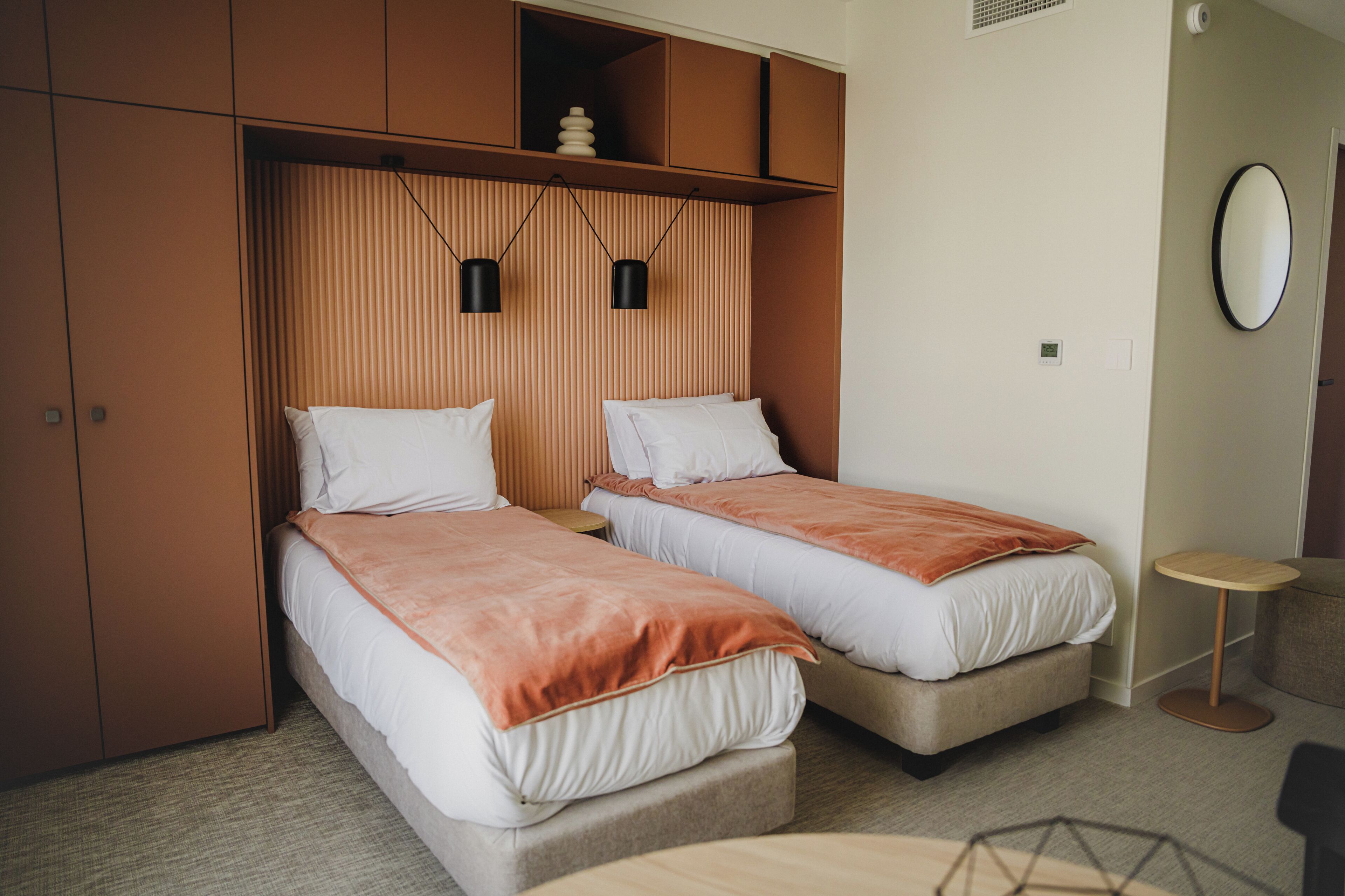 Une chambre d’hôtel avec deux lits, une petite table, un miroir rond et une armoire.