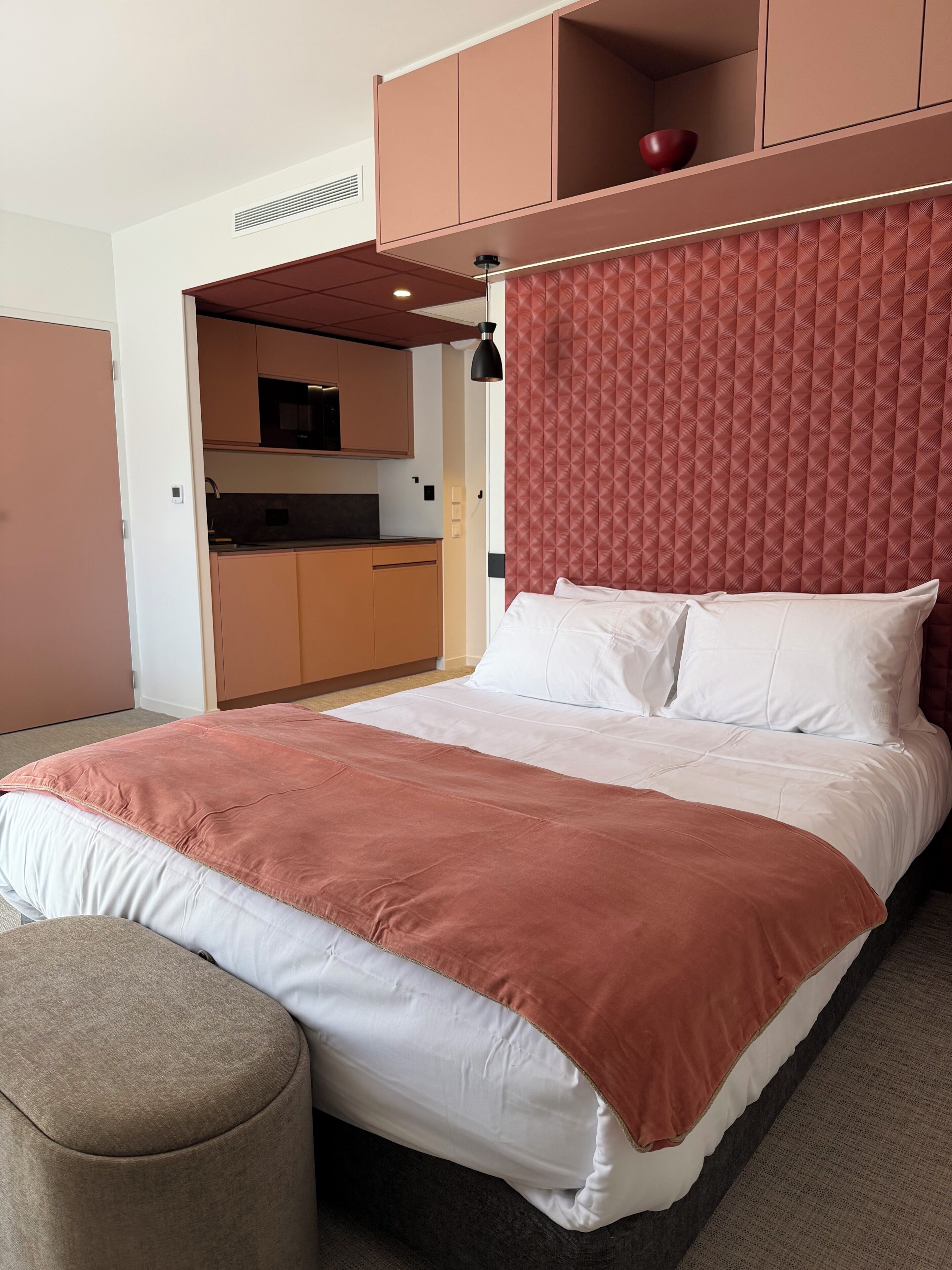 Une chambre à coucher avec un lit, une table de chevet, une tête de lit rouge et, au fond, une petite cuisine.