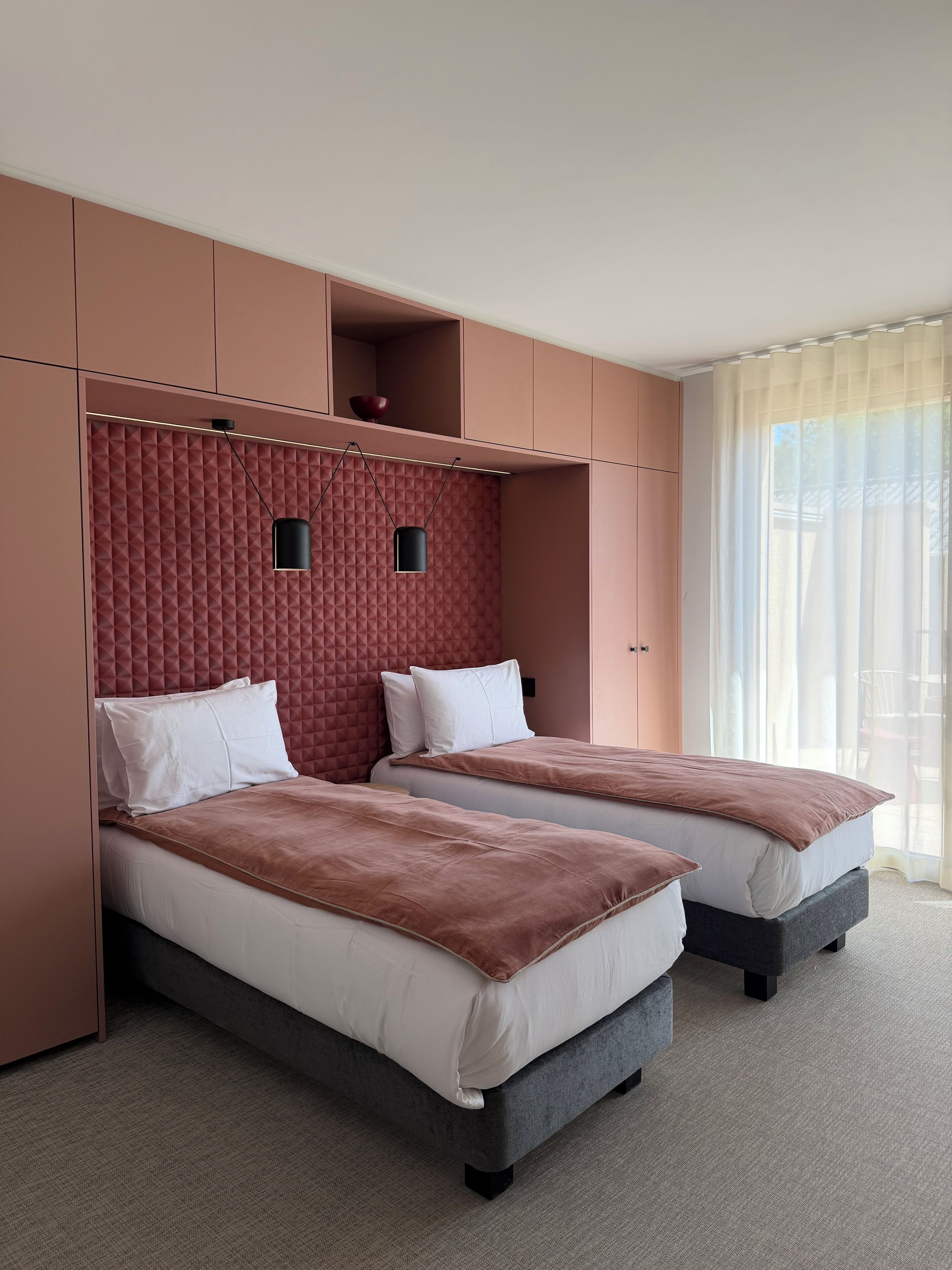 A hotel room with two beds, a built-in wardrobe, and a large window with sheer curtains.