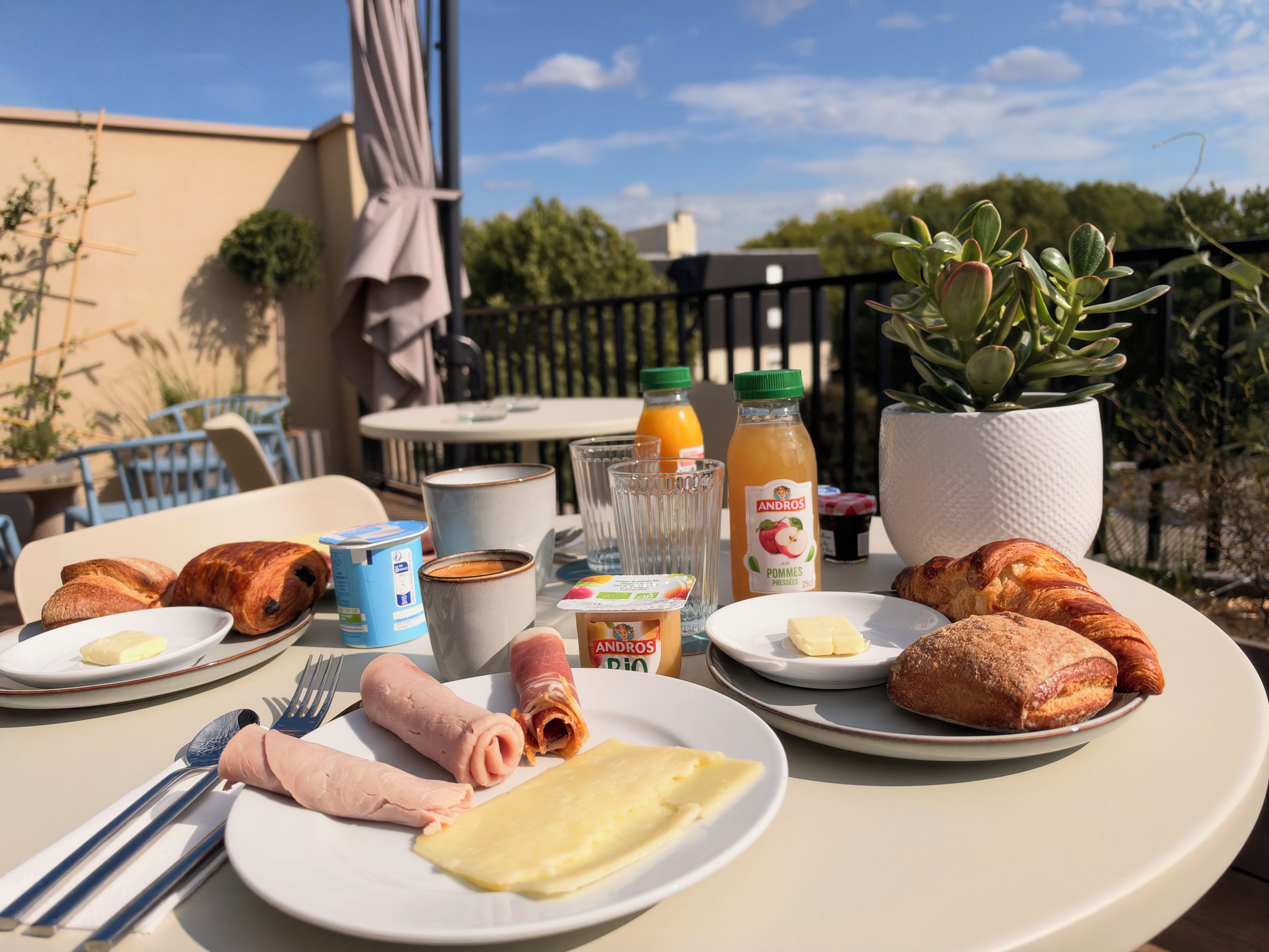 Ein Tisch ist reichhaltig mit Frühstück zubereitet, darunter Croissants, Schinken, Käse und Saft, mit Blick auf Grünflächen und ein Gebäude im Hintergrund.