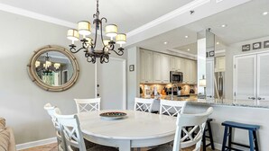 A dining area with a round table, white chairs, a chandelier, and a kitchen in the background. - Coastal Comfort at TOPSL Summit 309C (Miramar Beach)