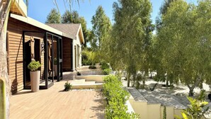 A modern building with wooden siding, a covered porch, and a landscaped garden with trees and shrubs. - Kenz Essia Luxury Countryside Escape Near Tunis (Jedaida)