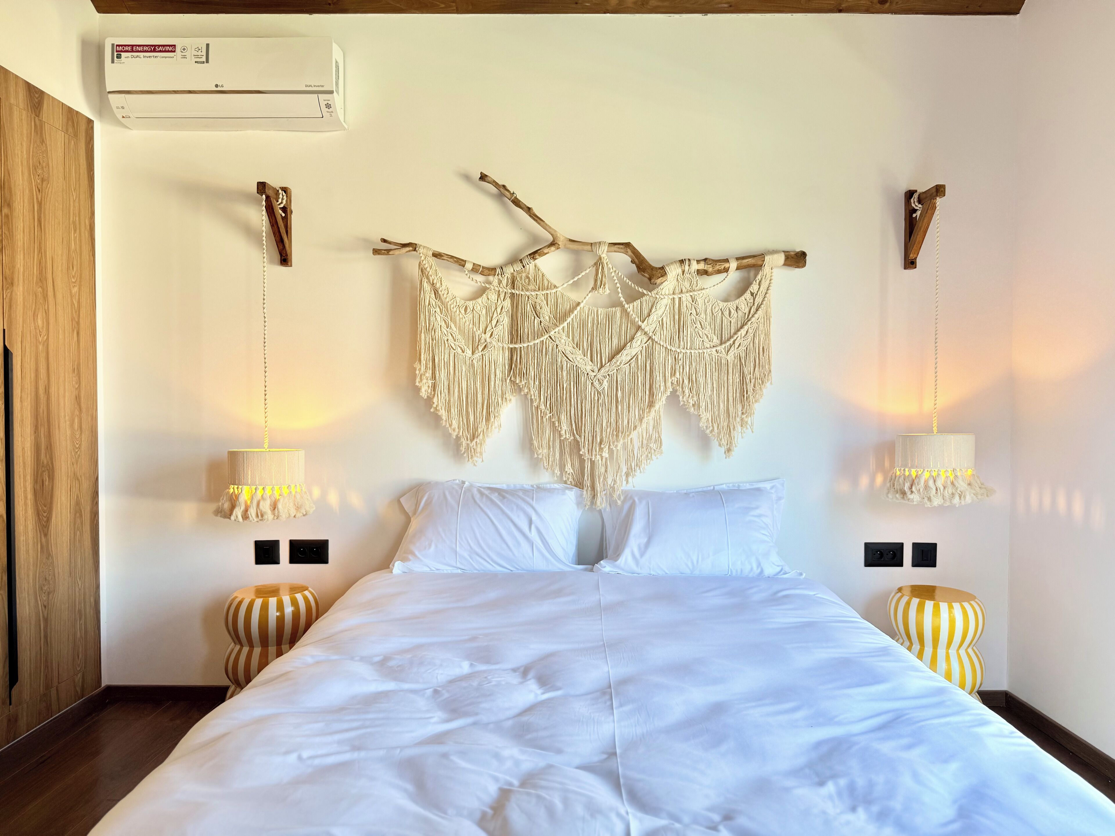 Ett hotellrum med en säng, en väggbonad i makramé, två sänglampor och en luftkonditioneringsanläggning.