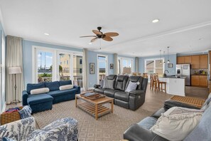 A spacious living area with a sofa, armchairs, and a coffee table. There is a ceiling fan, a kitchen area with wooden cabinets, and large windows offering a view. - Waterview 3BR | Steps to Beach with Pool (Carolina Beach)