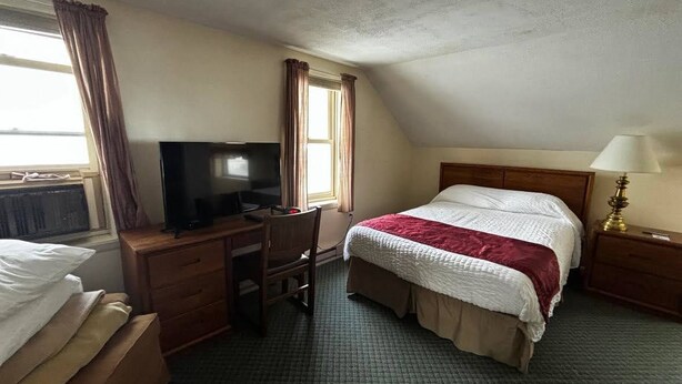 Quarto de hotel com cama, escrivaninha, televisão, luminária e janela com cortinas.