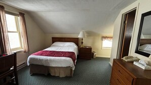 Quarto de hotel com uma cama de solteiro, mesa de cabeceira com abajur, cômoda de madeira e televisão.