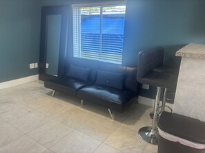 A room with a dark blue wall, a window with blinds, a black leather sofa, a small bar area with a stool, and a tiled floor. - Heart of Wynwood Spacious 2bedroom #3 (Miami)