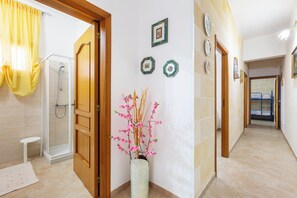 Un pasillo de hotel con ducha, una puerta de madera, un jarrón con flores rosadas y una cama a lo lejos.