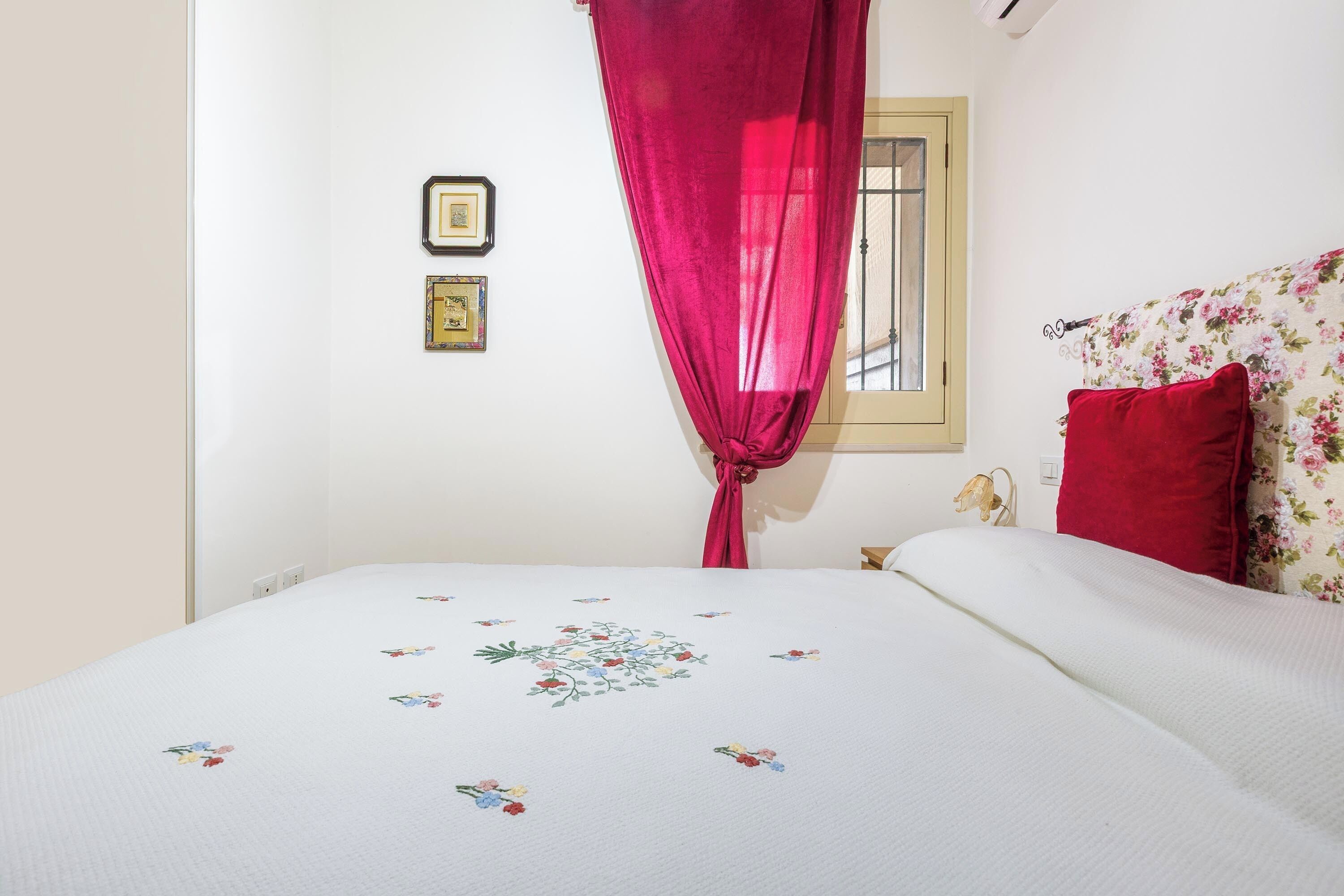 A bedroom with a bed, a window with curtains, a wall-mounted light fixture, and two framed pictures.