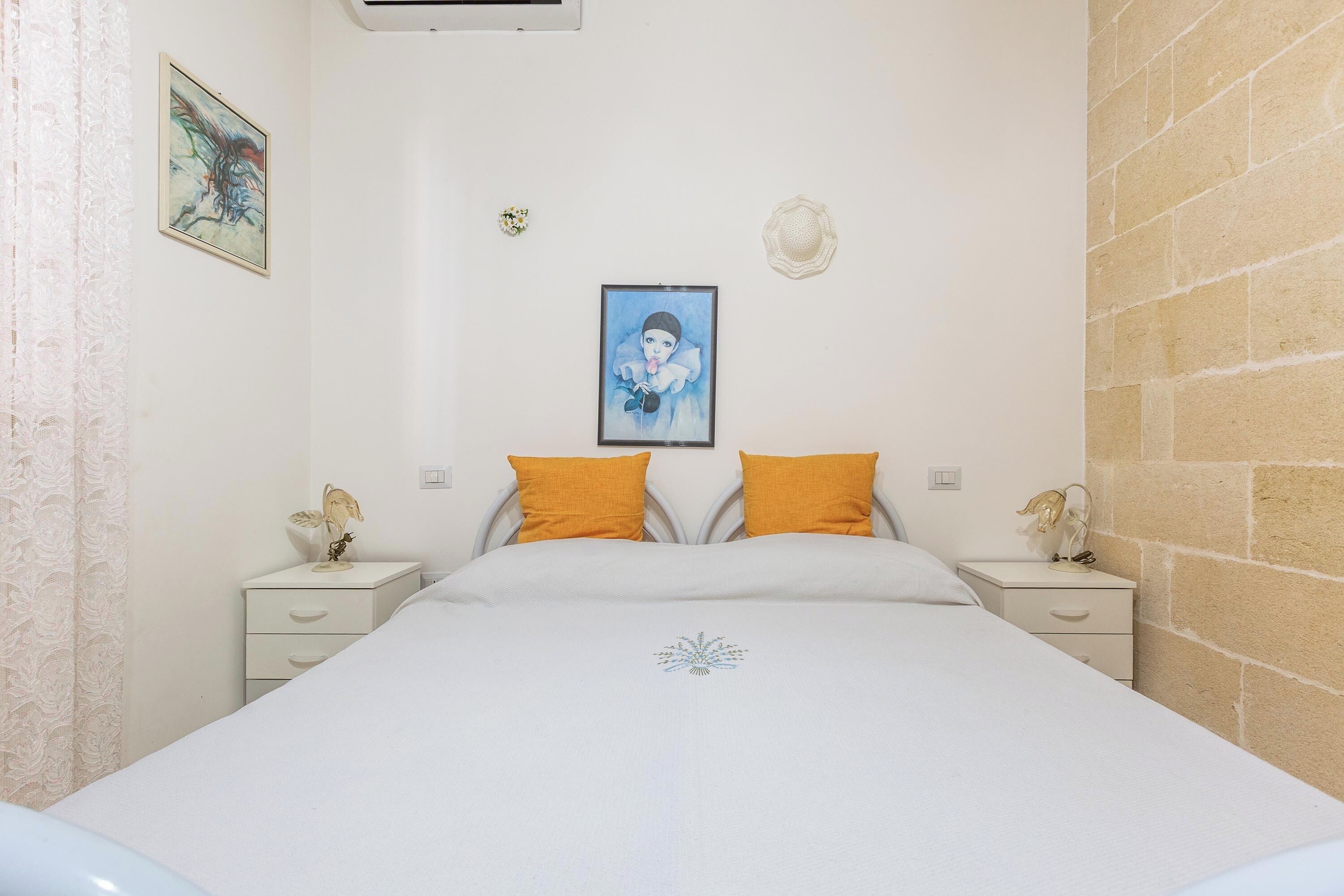 A bedroom with a bed, two nightstands, a wall-mounted air conditioner, and a single artwork.