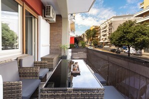 A balcony with wicker furniture and a glass table, overlooking a street with buildings and parked cars. - Due Passi dal Vaticano by BarbarHouse (Roma)