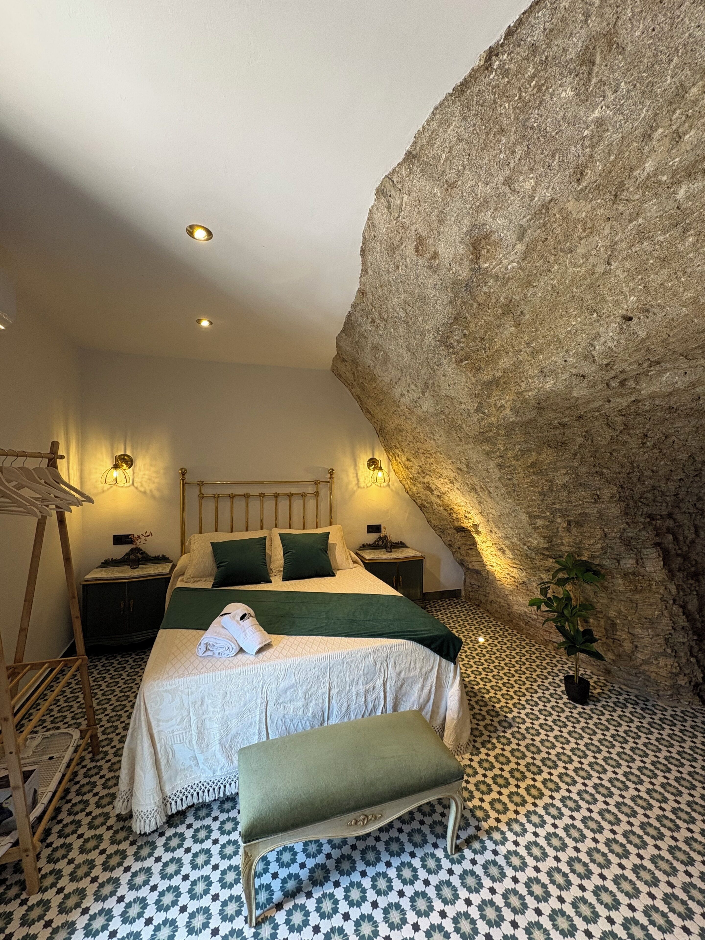 A bedroom with a large rock wall, a bed with green bedding, a bench, and a potted plant.