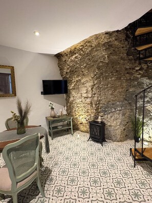 A room with a stone wall, a flat-screen TV mounted on the wall, a dining table with chairs, and a patterned floor. - The Jewel of Setenil - Esmeralda Cave House (Setenil de las Bodegas)