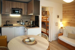 Ett litet kök med mikrovågsugn, diskho och träskåp. Där finns ett matbord med två skålar och en stol. I det intilliggande rummet syns en våningssäng med ett draperi.