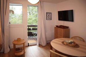 Ein Zimmer mit Holztisch, Stühlen, einem Fernseher, einem Korbschrank und einem Balkon mit einem Sessel.
