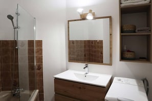 Ein Badezimmer mit einer Duschkabine aus Glas, einem weißen Waschbecken mit Holzunterschrank, einem spiegel mit Holzrahmen sowie einem Regal mit Handtüchern und Körben.