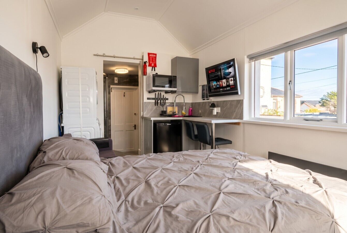 Un dormitorio compacto con cama, kitchenette, televisión y ventana.