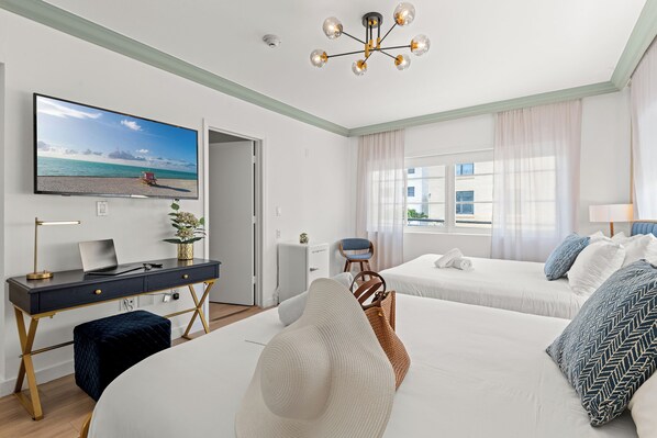 Une chambre d’hôtel moderne avec un grand lit, un bureau avec un ordinateur portable, un téléviseur diffusant une scène de plage et une vue sur la mer par la fenêtre.