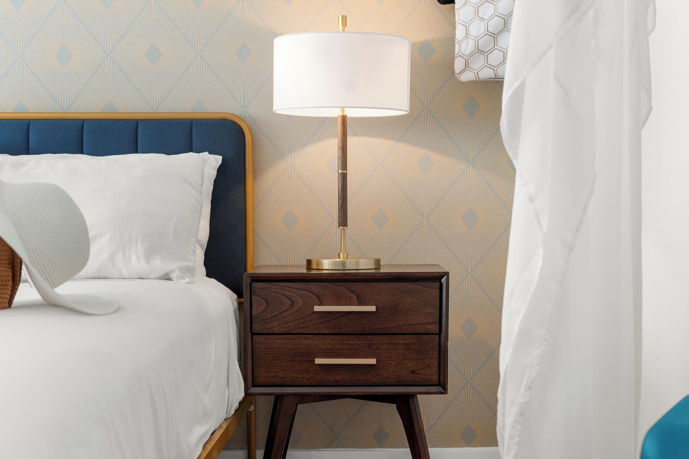 A neatly made bed with white bedding and a blue headboard, a bedside table with a lamp, and a patterned wallpaper.
