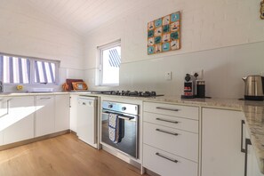 A kitchen with white cabinets, a stove, a coffee maker, and a window with blinds. - Tranquil 3-bedroom house in charming Onrusrivier for your getaway (Onrusrivier)