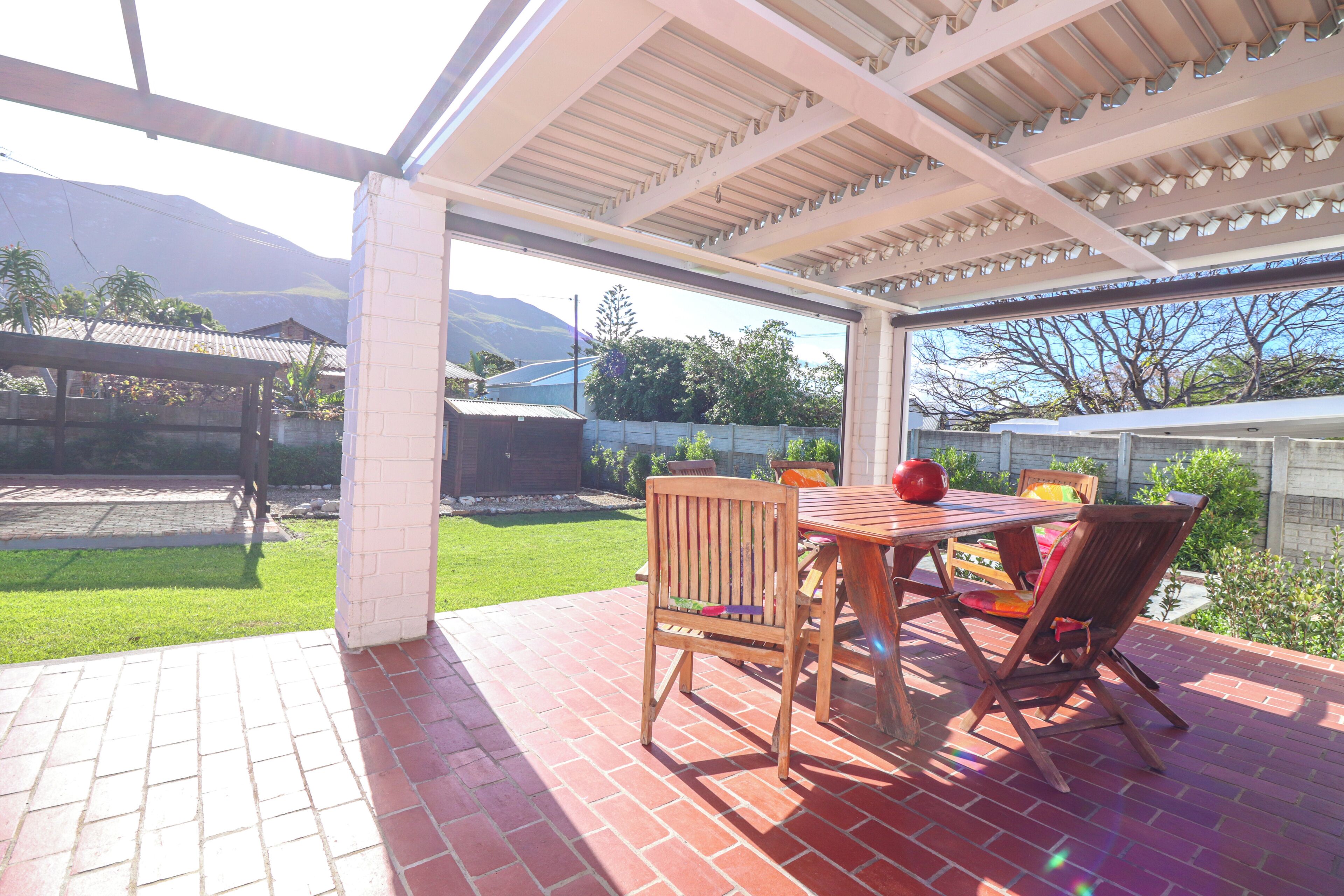 A covered patio with a brick floor, wooden furniture, and a view of a backyard with a wooden fence and greenery.
