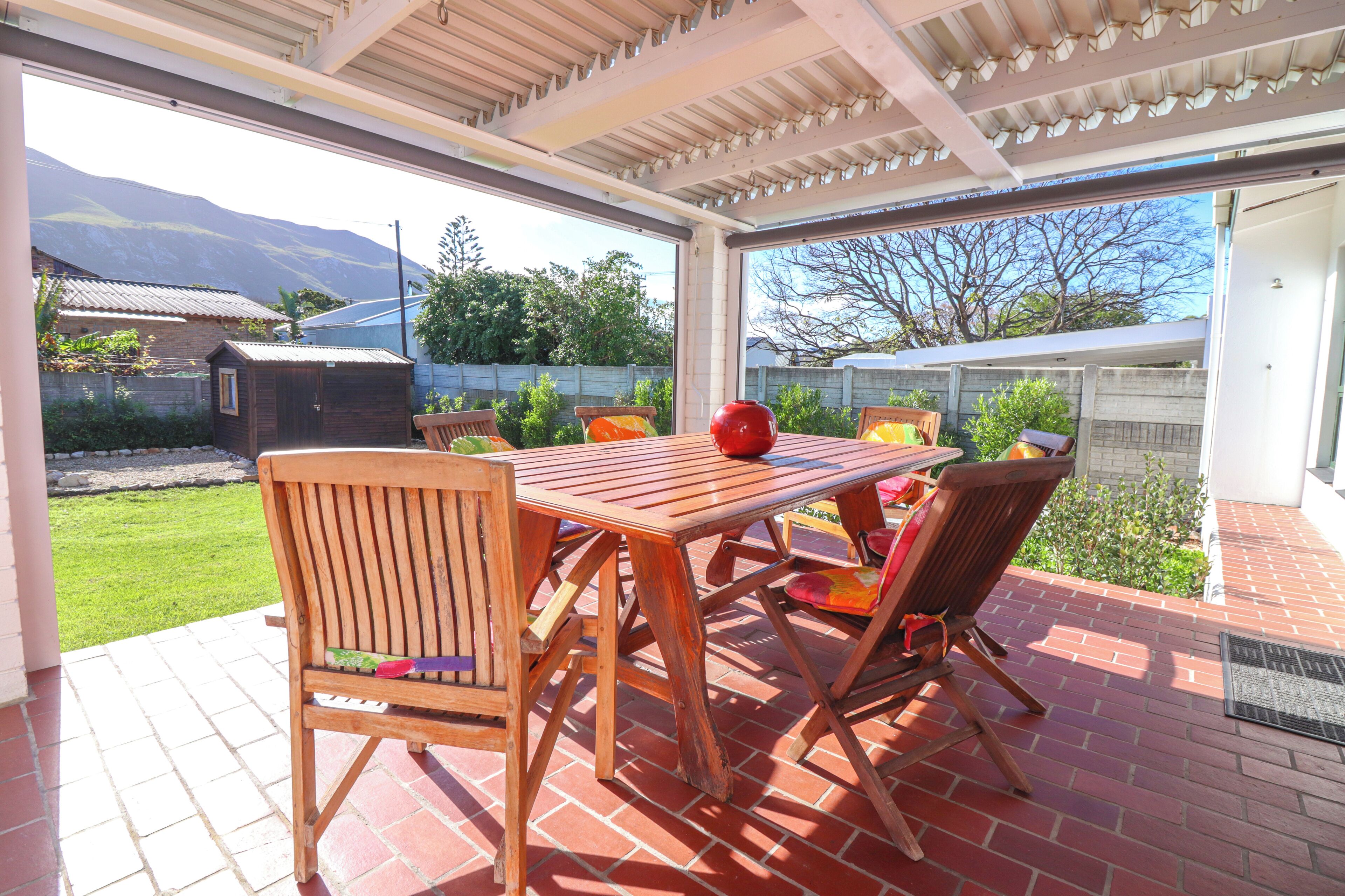 An outdoor patio with a wooden table and chairs, a red vase, and a view of a mountain and garden.