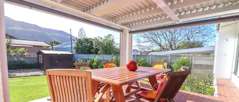 Un patio al aire libre con una mesa y sillas de madera, un florero rojo y vistas a una montaña y un jardín.