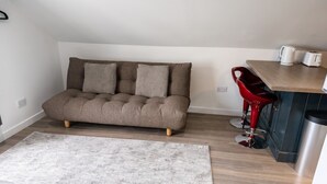 Un espace de vie compact comprenant un canapé, un coin repas avec une petite table et des chaises, et un coin cuisine avec un évier et une bouilloire.