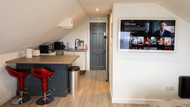 Une cuisine moderne avec un coin bar, un téléviseur fixé au mur diffusant un service de streaming, et une porte donnant sur une autre pièce.