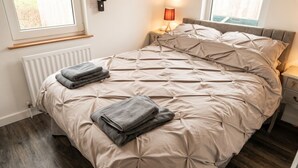 Een net opgemaakt bed met een beige dekbed en kussens, een bedlampje en een raam met uitzicht op groen.