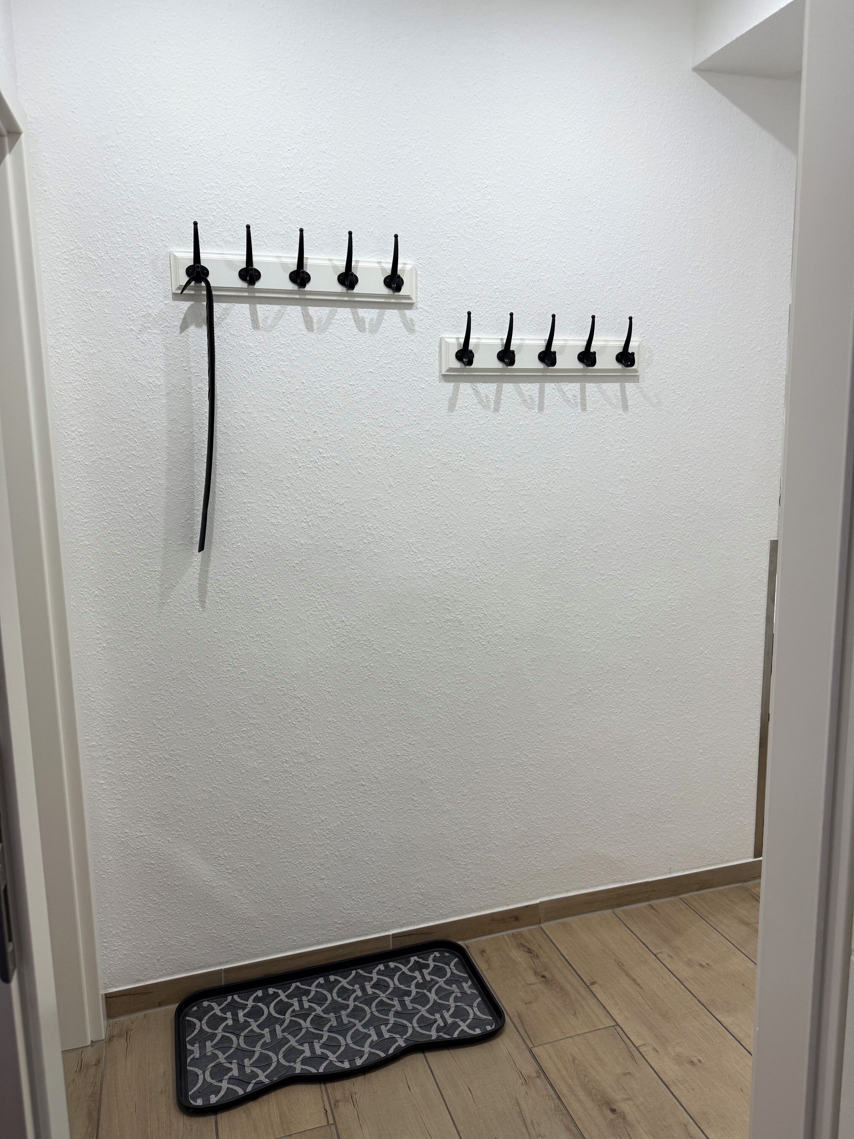 A hallway with a white textured wall, a black doormat with a geometric pattern, and two wall-mounted coat hooks.