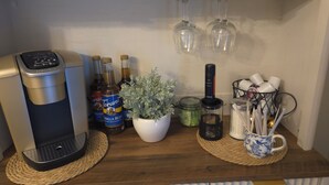 Pieni keittiönurkkaus, jossa on kahvinkeitin, Torani-siirappipulloja, Keurig-kahvinkeitin, ruukkukasvi, kahvimylly ja muki, jossa on aterimet.