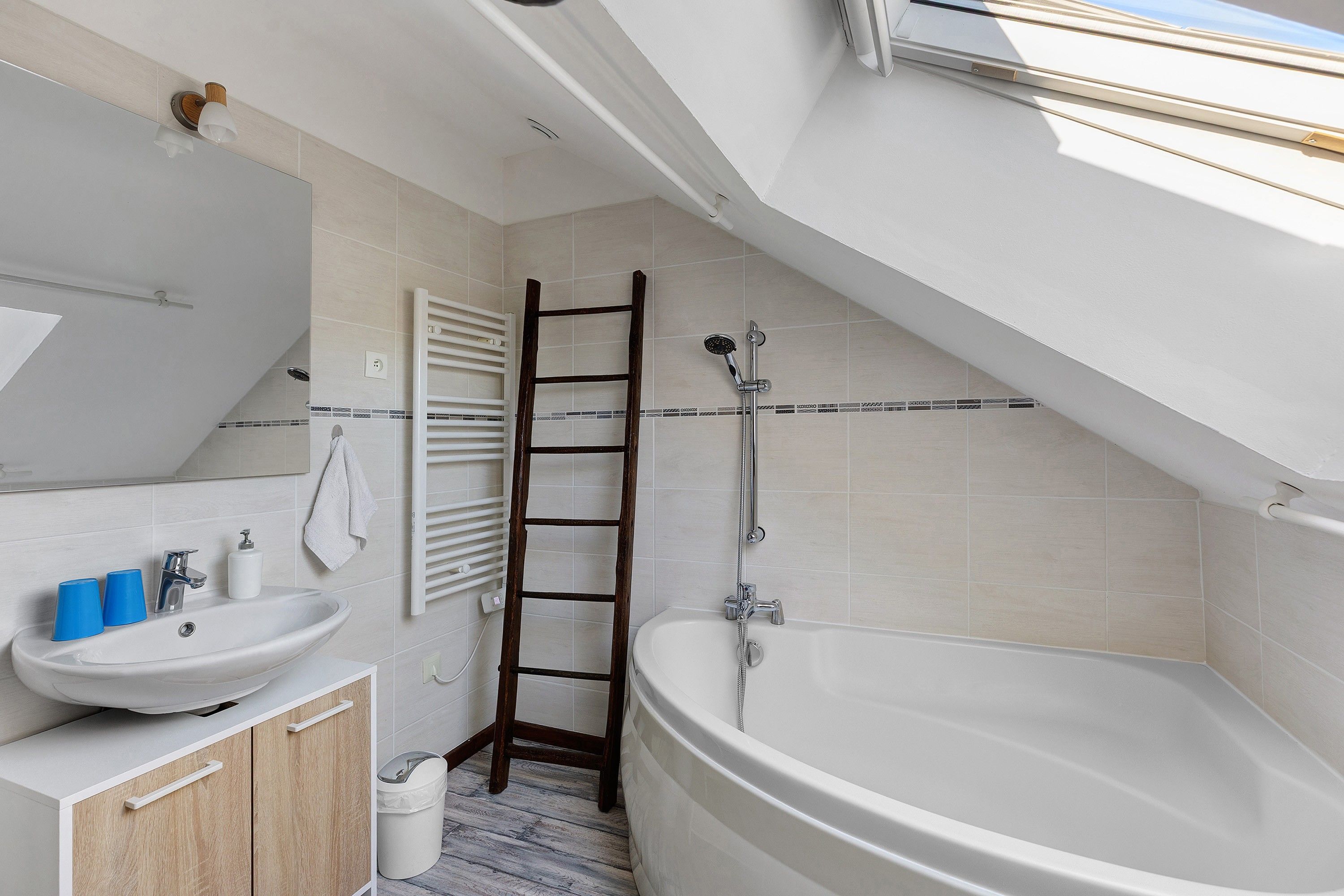 A modern bathroom with a freestanding bathtub, a wooden ladder, a white sink with blue cups, and a large mirror.