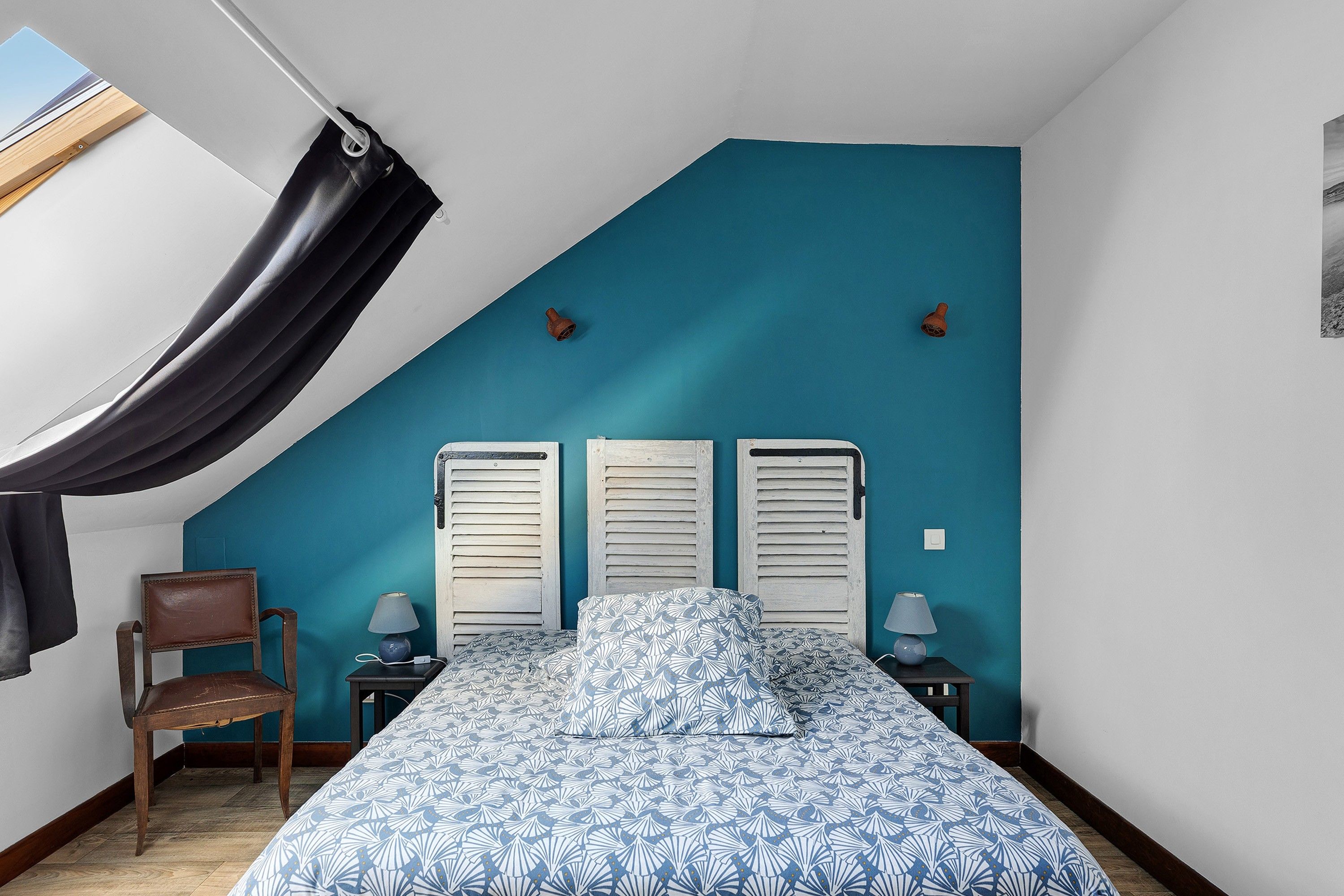 Ein Schlafzimmer mit geneigter Decke, einem Bett mit gemusterten Betttextilien, einem Sessel, zwei Nachttischen mit Lampen und einem Fenster mit Vorhang.