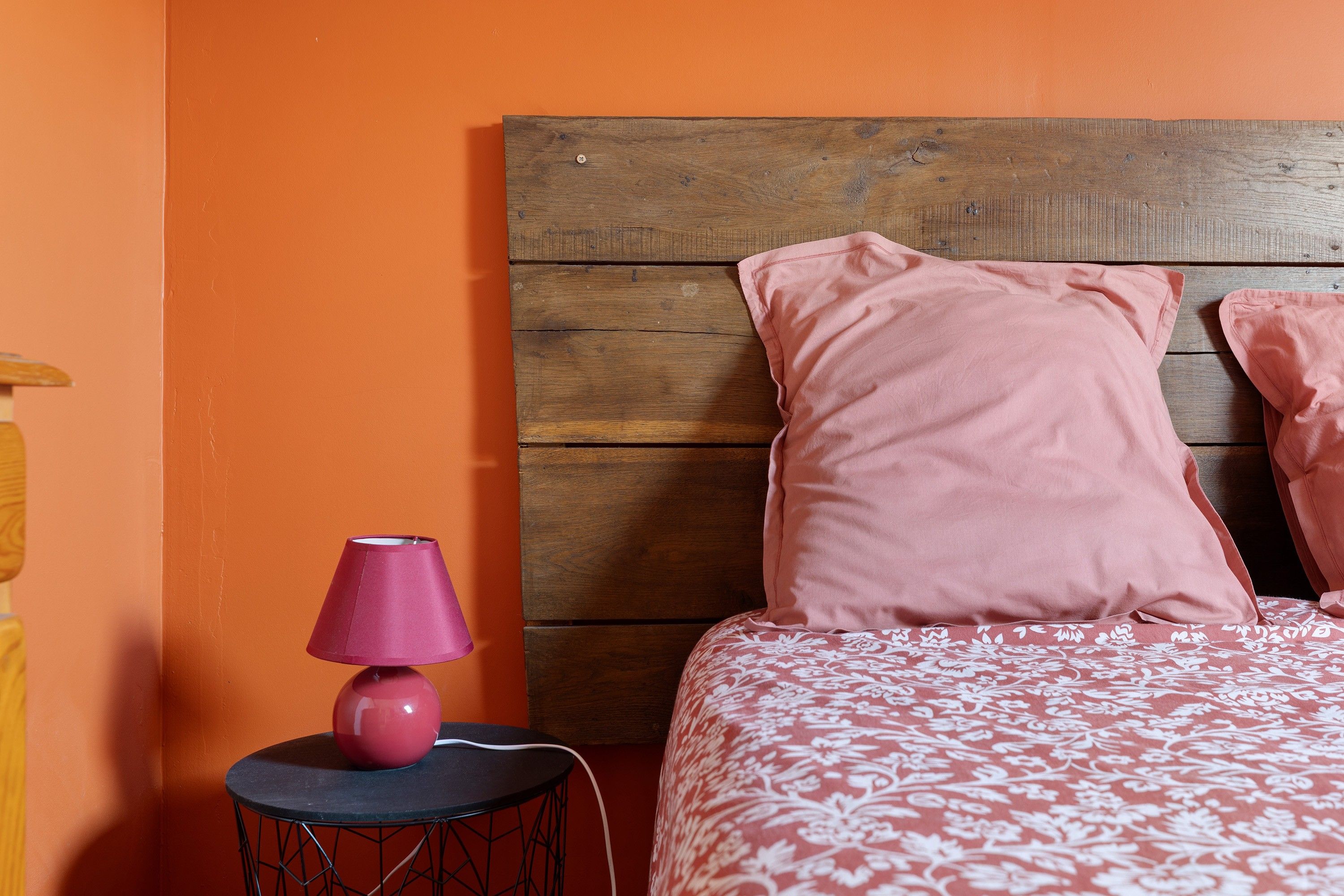 Een slaapkamer met een houten hoofdeinde, een roze lamp op een klein tafeltje en een bed met roze beddengoed.