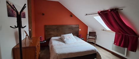 A bedroom with a sloped ceiling, a bed with a wooden headboard, a nightstand, a chair, a coat rack, and a window with curtains.