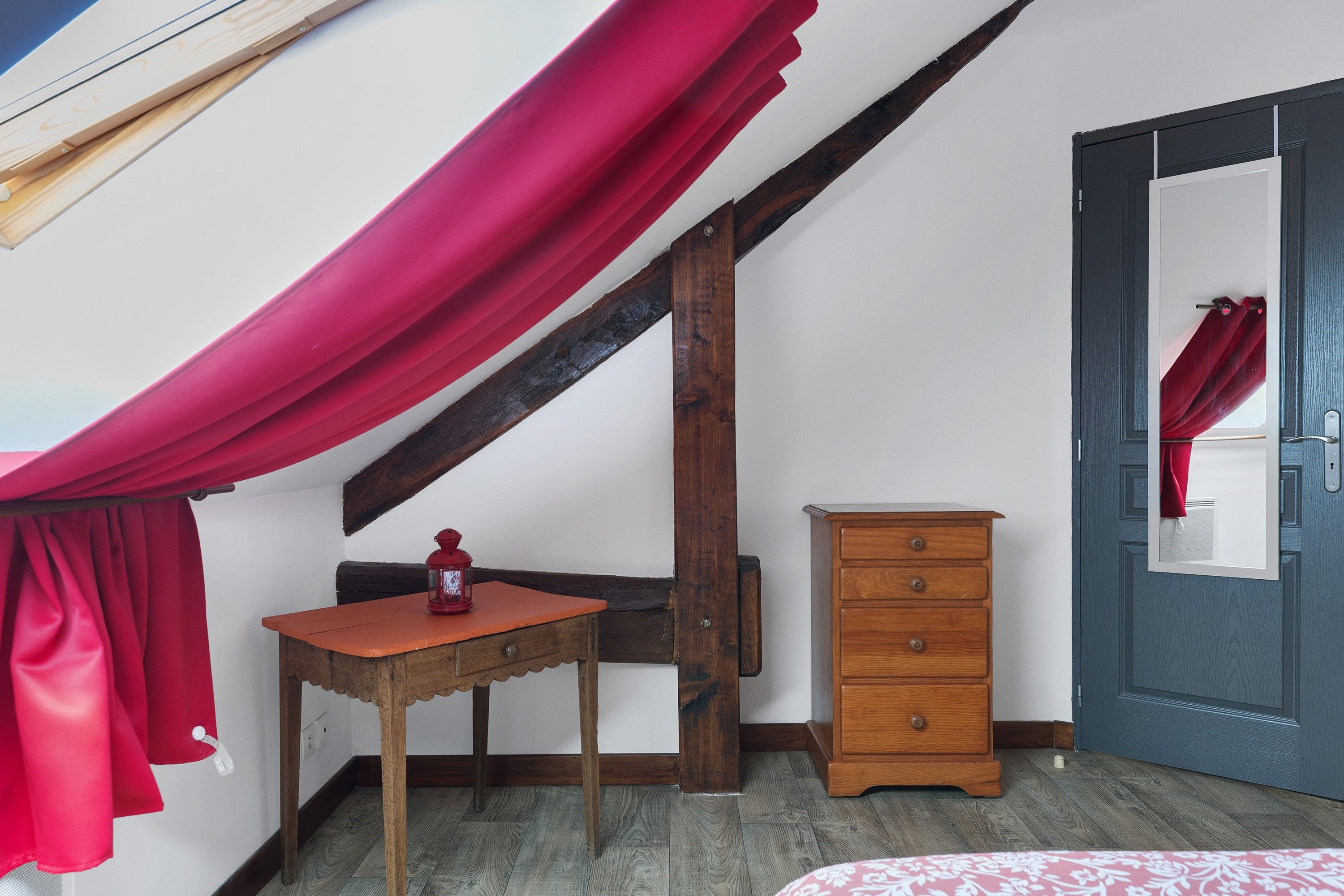 A room with a sloped ceiling, a wooden table with a red lantern, a wooden dresser, and a door with a red curtain.
