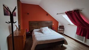 Un dormitorio con techo inclinado, una cama con ropa de cama estampada, un aparador de madera, una silla y un ventanal con cortinas rojas.