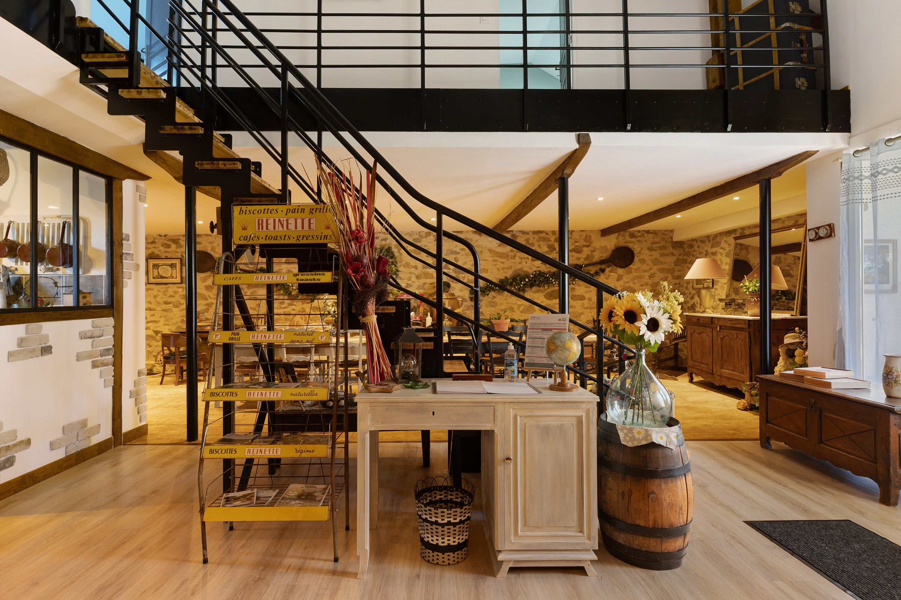 Un escalier avec une rampe noire menant à un deuxième étage, une table en bois ornée d’un globe terrestre et de fleurs, et une enseigne indiquant les produits de la boulangerie « REINETTE ».