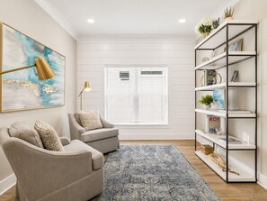 Ein Wohnzimmer mit zwei Sesseln, einem blauen Teppich mit Muster, einem Bücherregal und einem großen abstrakten Gemälde.