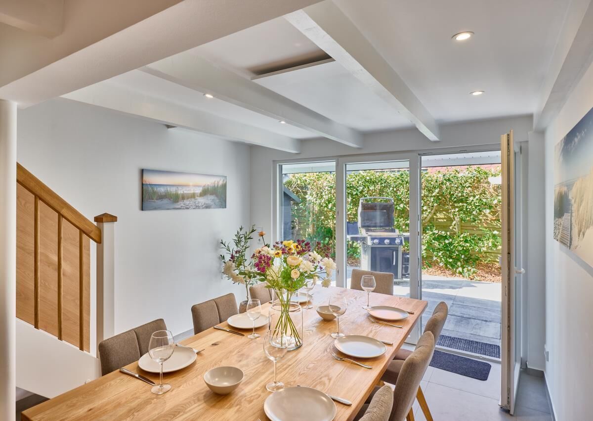 Un coin repas avec une table en bois dressée pour quatre personnes, entourée de chaises, et donnant sur un barbecue situé à l’extérieur.