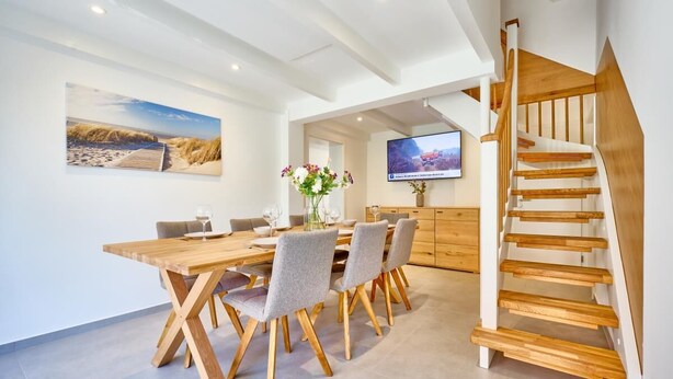 A modern dining area with a wooden table and chairs, a staircase, and a flat-screen TV mounted on the wall. - Vacation Home at Am Ringwal 82 in Cuxhaven Duhnen—modernized in 2026 with a Wallbox (Cuxhaven)