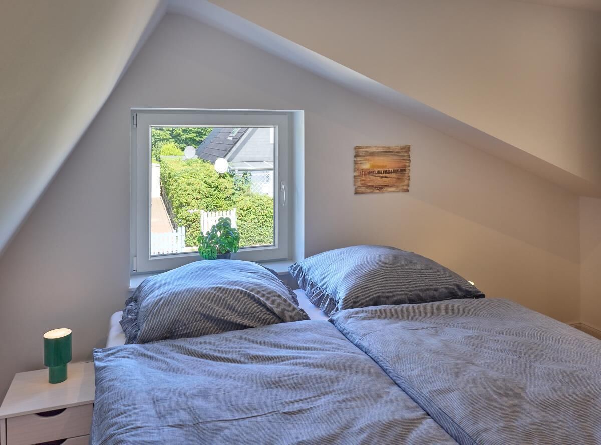 A bedroom with a bed, a nightstand, a lamp, a window, and a wall-mounted artwork.