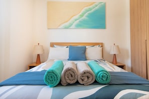 Ein ordentlich bezogenes Bett mit aufgerollten Handtüchern, zwei Nachttischlampen und einem Bild an der Wand.