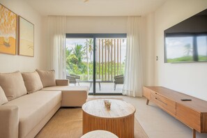 A modern living room with a sofa, a coffee table, and a TV. There is a sliding glass door leading to a balcony with outdoor furniture. - Born | Family Getaway with Pool View (Bavaro)