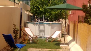 Ein kleiner Hinterhof mit einem Pool, zwei Liegestühlen, einem blauen Stuhl, einer Leiter und einem grünen Sonnenschirm.