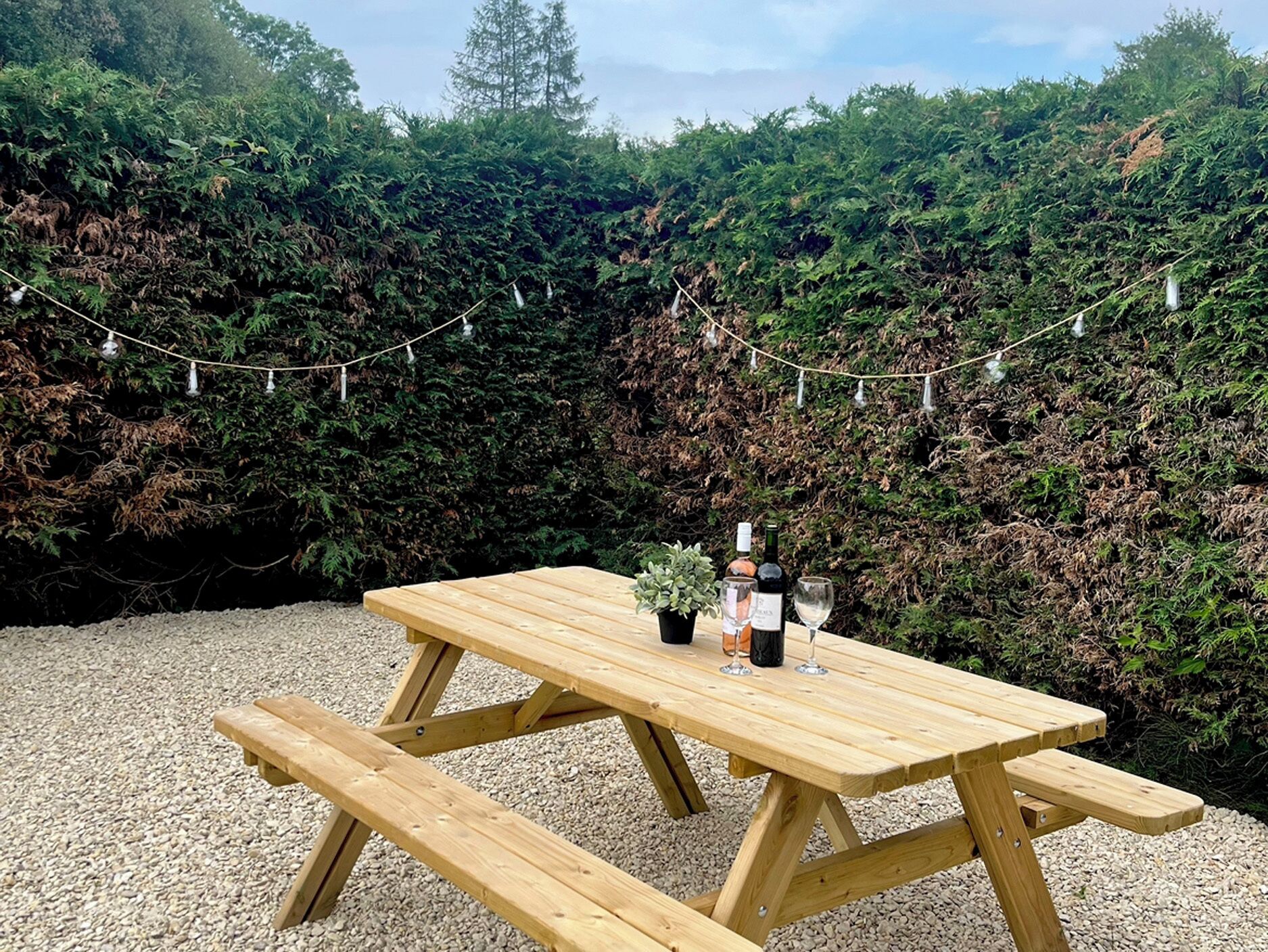 Een houten picknicktafel met bankjes, een fles wijn en een glas erop, geplaatst op grind met sfeerverlichting op de achtergrond.