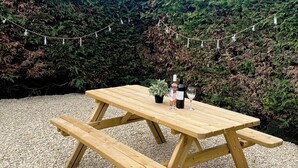 Ein Holztisch mit Bänken, eine Weinflasche und ein Glas darauf, auf Kies, im Hintergrund Lichterketten.