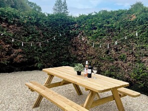 Ein Holztisch mit Bänken, eine Weinflasche und ein Glas darauf, auf Kies, im Hintergrund Lichterketten.