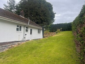A white house with a tiled roof, a front door, and a small front yard with grass and a hedge. - Saddleback Bungalow (Llandrindod Wells)
